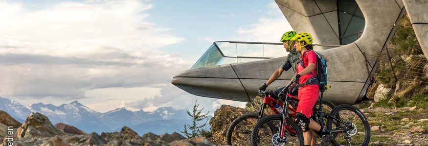 Zwei Radfahrer in Bergkleidung vor einer futuristischen Bergstation mit Alpenblick