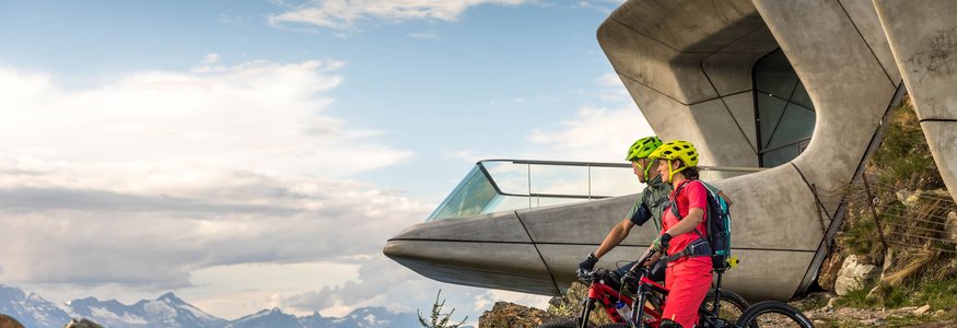 Mountainbike-Saisonstart am Kronplatz © WOM Medien - Andreas Meyer Zwei Radfahrer in Bergkleidung vor einer futuristischen Bergstation mit Alpenblick