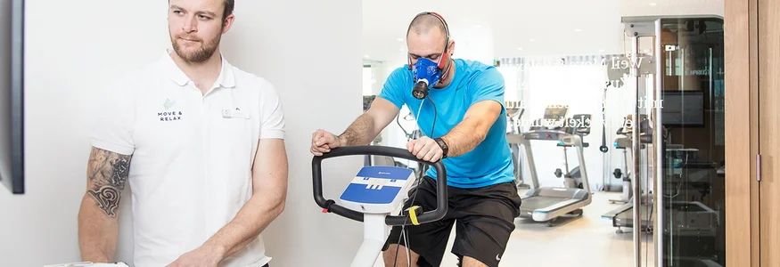 Mann mit Atemmaske auf Fahrradtrainer wird von Trainer überwacht
