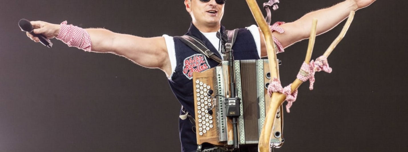 Man on stage with accordion and microphone, arms spread wide