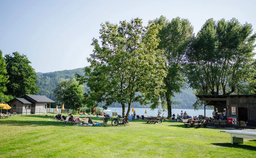 Grüner Park mit Bäumen, Liegestühlen und Menschen am Seeufer bei sonnigem Wetter