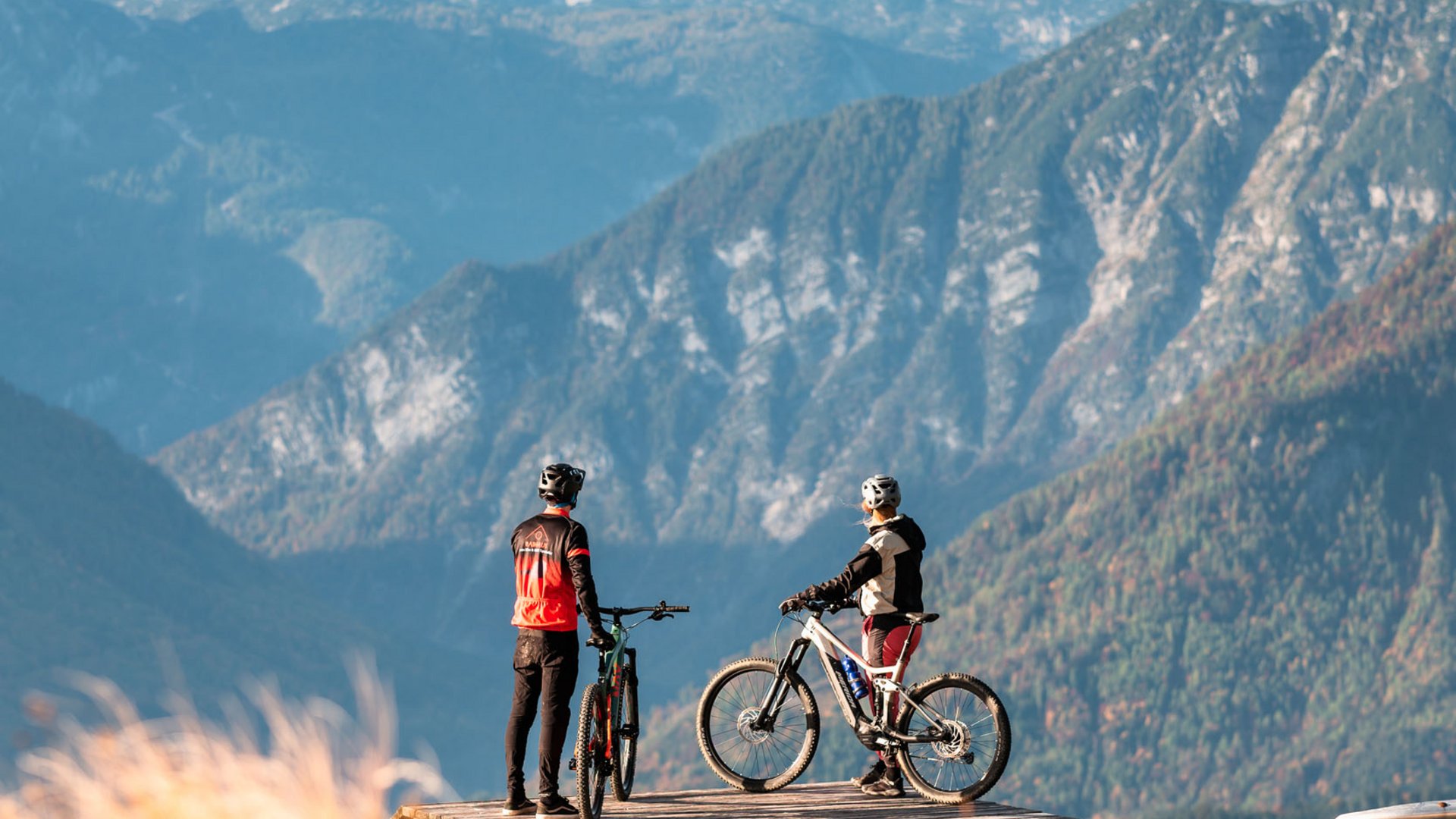 Zwei Mountainbiker auf Holzplattform mit Blick auf schneebedeckte Berge