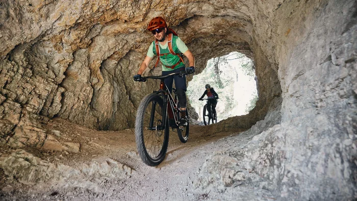 Zwei Mountainbiker fahren durch einen Felstunnel auf einem schmalen Pfad