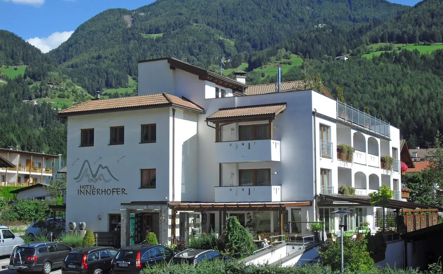 Hotel Innerhofer with forested mountains and parked cars under blue sky
