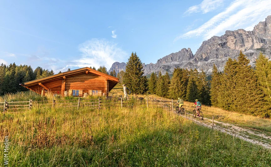 Two cyclists on a path near a wooden cabin with mountains in the background