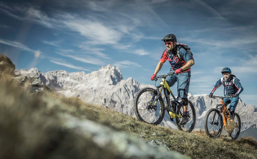 Zwei Mountainbiker fahren bergauf mit Bergkulisse und blauem Himmel