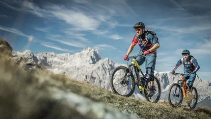 Zwei Mountainbiker fahren bergauf mit Bergkulisse und blauem Himmel