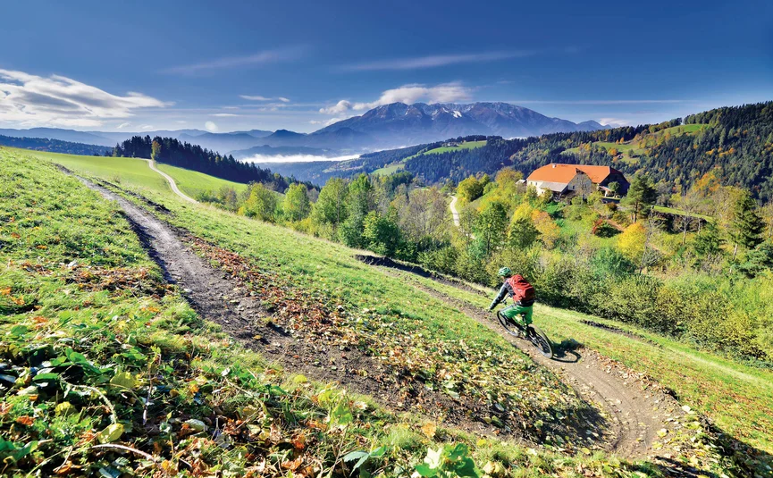 Mountainbiker auf Trail in grüner Hügellandschaft mit Berg und Haus