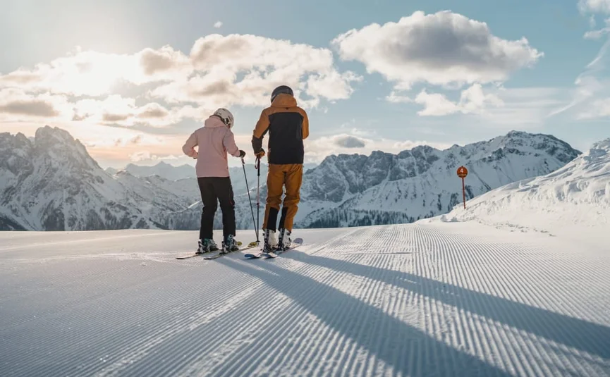Zwei Skifahrer auf einer präparierten Piste vor verschneiten Bergen