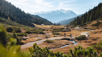 Two cyclists on a mountain trail with snow-capped peaks in the background