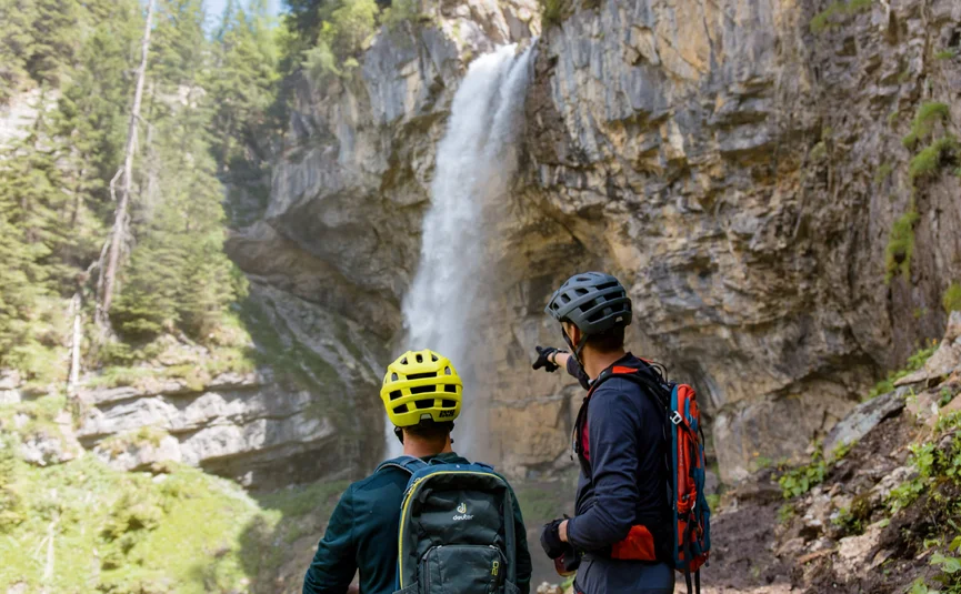 Zwei Radfahrer in Helmen betrachten einen Wasserfall in einer felsigen Landschaft