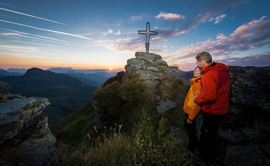 Koppel in wandeljassen op bergtop naast kruiskruis bij zonsondergang