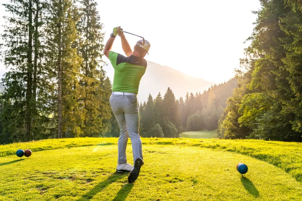 Golfer hitting ball on sunny golf course with forest and mountains behind