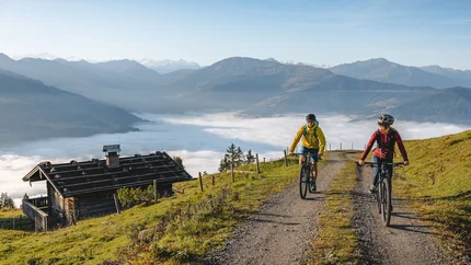 Zwei Radfahrer fahren auf Bergweg mit Nebel im Tal und Bergen im Hintergrund