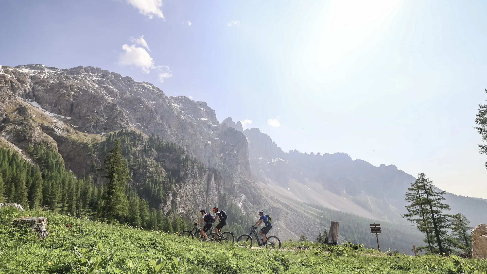 Genuss-(E-)Biken in den Dolomiten © Jens Vögele - Westsiders Drei Mountainbiker fahren im Gebirge auf grünem Pfad bei sonnigem Wetter