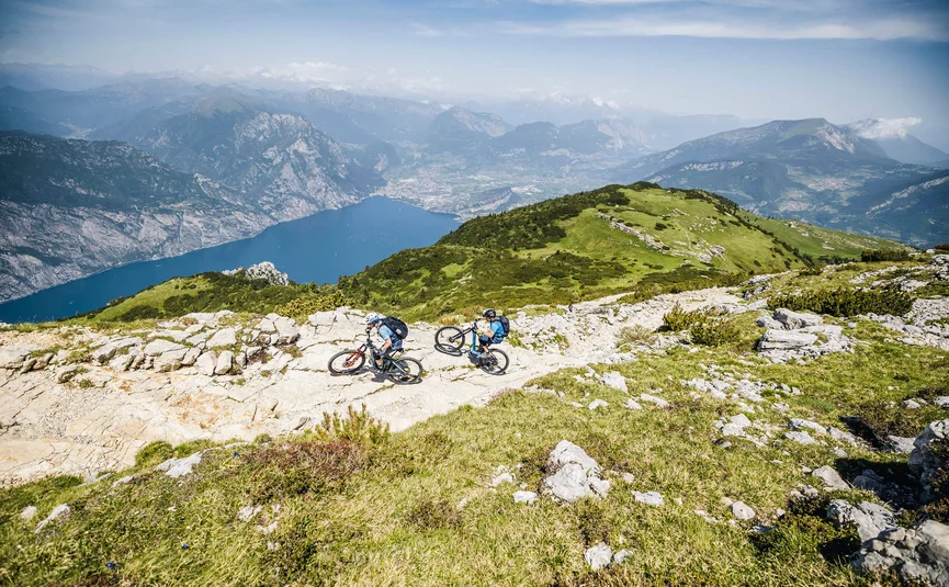 Two mountain bikers on rocky trail with lake and mountains in background