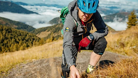 Bikeurlaub per Bahn © David Karg Mann mit Helm und Handschuhen untersucht Pflanzen auf Bergwanderweg