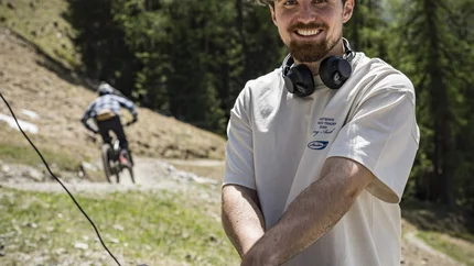 Smiling DJ with headphones outdoors near trees and a cyclist in the background