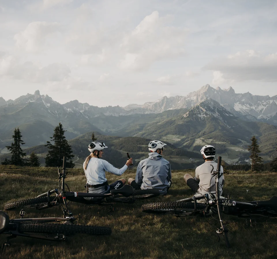 Drei Radfahrer sitzen auf einer Wiese mit Blick auf Berge und tragen Helme