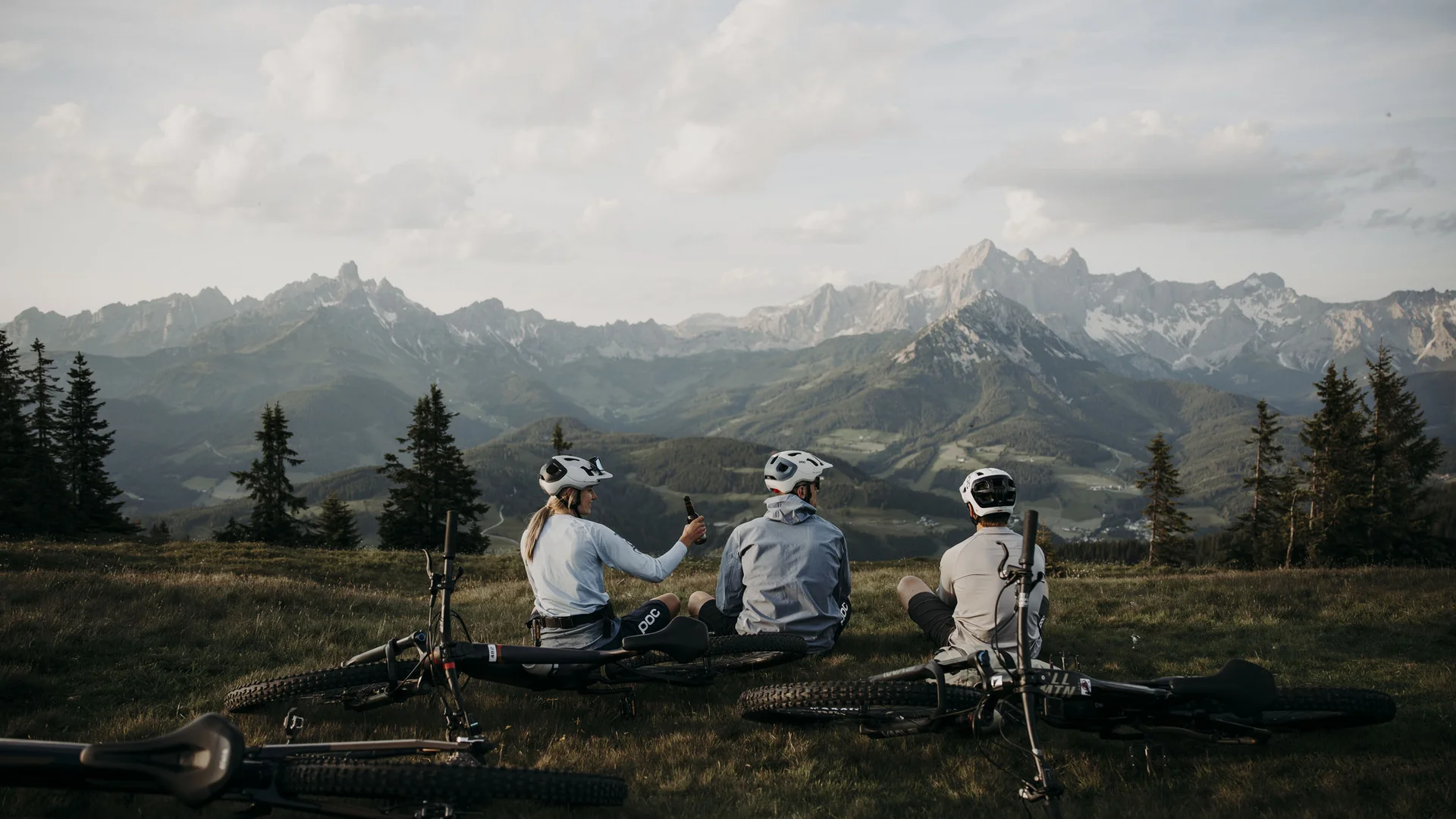 Bikeurlaub per Bahn © Matthias Warter Drei Radfahrer sitzen auf einer Wiese mit Blick auf Berge und tragen Helme