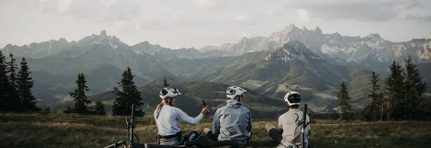 Drei Radfahrer sitzen auf einer Wiese mit Blick auf Berge und tragen Helme
