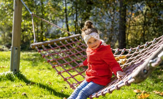 Brennseehof Happy Family Woche Mädchen in roter Jacke sitzt auf einem Netz-Hängematte im Garten