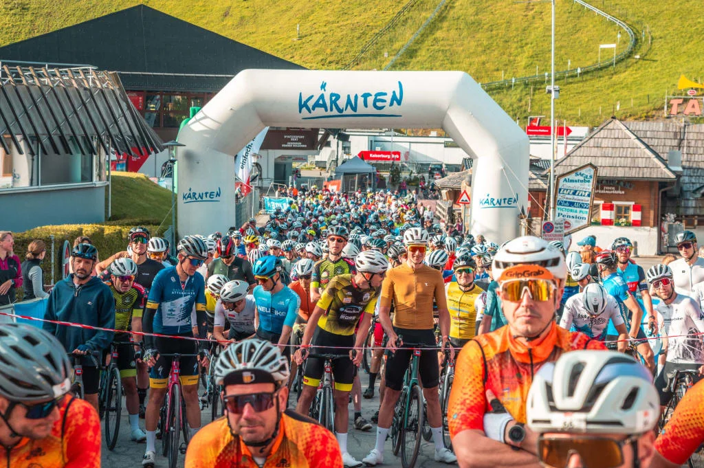 Start eines Radrennens mit vielen Fahrern unter einem Tor mit Schriftzug Kärnten