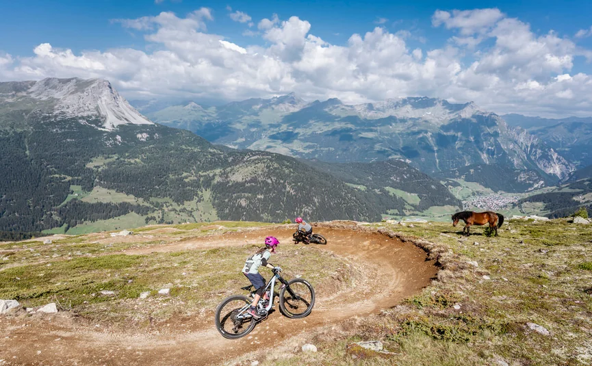 Zwei Kinder fahren Mountainbike auf Bergpfad mit Pferd und Alpen im Hintergrund