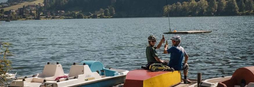 BEST OF Familien-Bikeurlaub © David Karg Zwei Männer auf Steg trinken Bier mit See und Bergen im Hintergrund