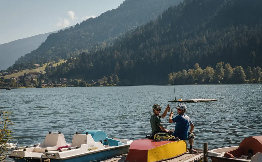 Two men on dock drinking beer with lake and mountains behind