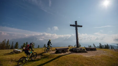 Zwei Mountainbiker am Gipfelkreuz bei sonnigem Bergwetter