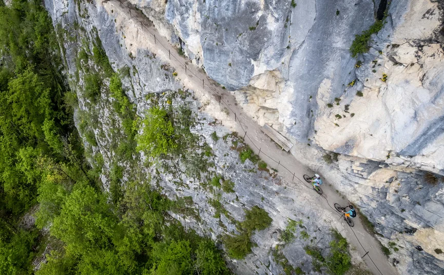Zwei Radfahrer auf schmalem Bergpfad an Felswand mit grünem Wald