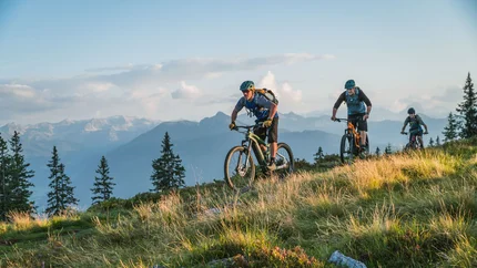 Drei Mountainbiker fahren auf einem grasigen Hügel mit Bergen im Hintergrund