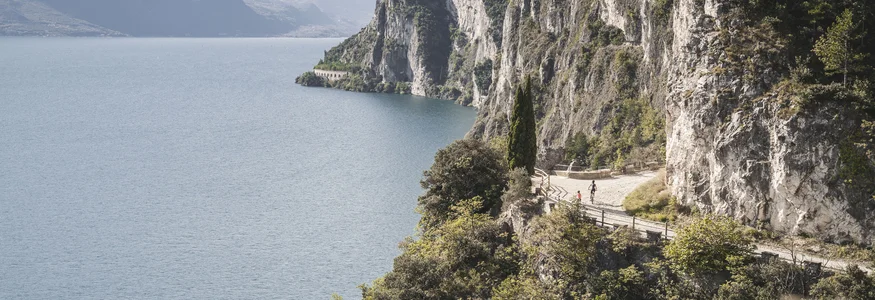 Cyclists on a mountain path by a lake with steep cliffs and forest