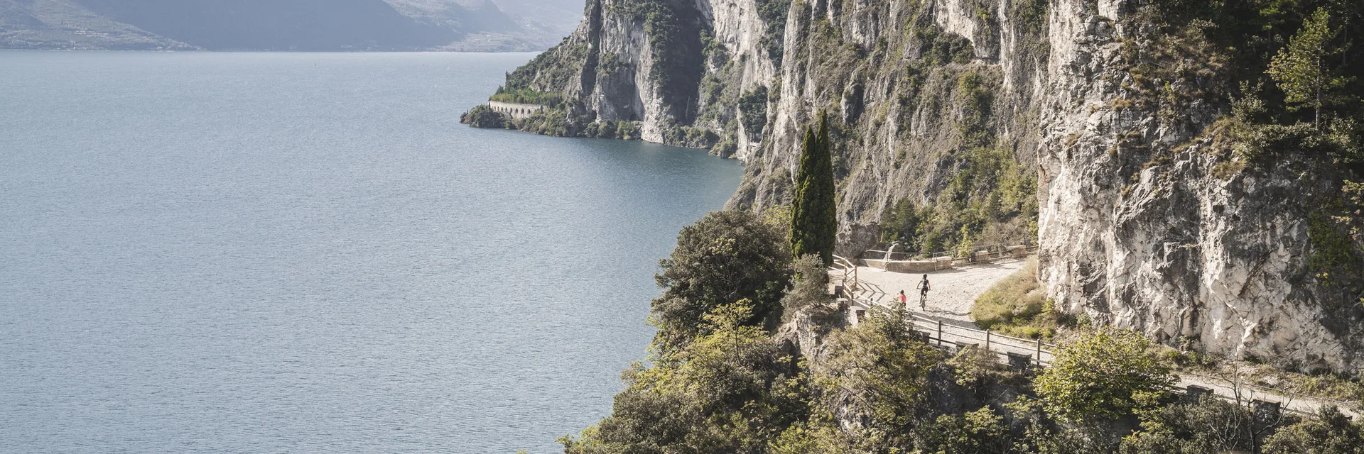 Cyclists on a mountain path by a lake with steep cliffs and forest