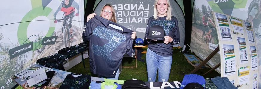 GREEN DAYS 2026 in Nauders-Reschensee
© TVB-Tiroler-Oberland-Nauders-Henri-Lesewitz Zwei Frauen präsentieren Merchandise-Artikel auf einem 3 Länder Enduro Trails Stand