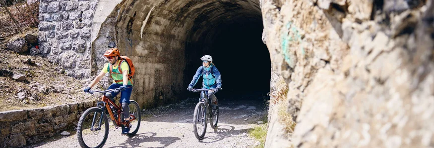 Zwei Radfahrer kommen aus einem Steintunnel auf einem Bergweg heraus