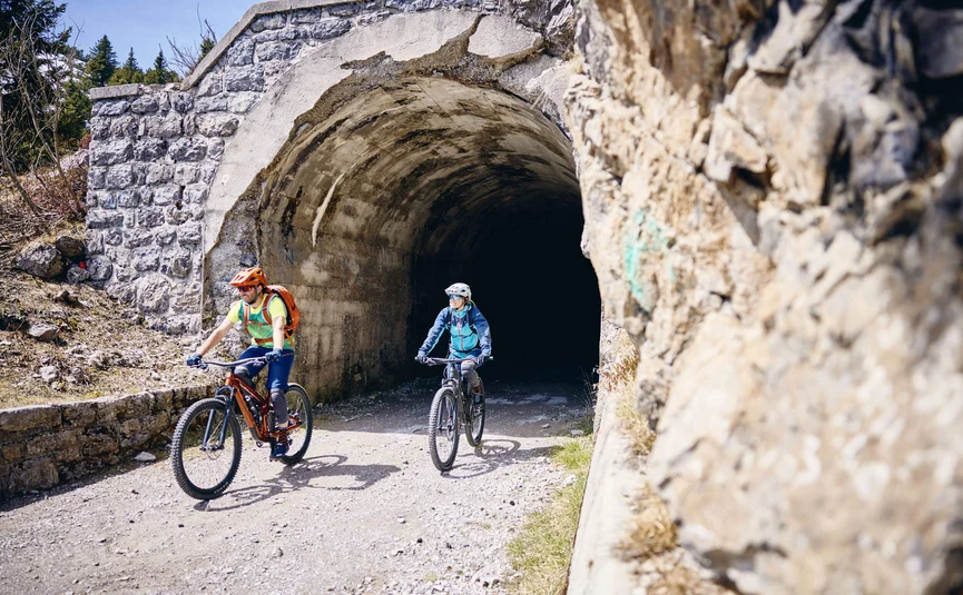 Zwei Radfahrer kommen aus einem Steintunnel auf einem Bergweg heraus