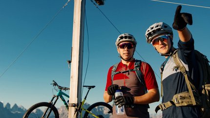 Zwei Radfahrer mit Helm und Rucksack am Gipfelkreuz mit Fahrrad im Hintergrund