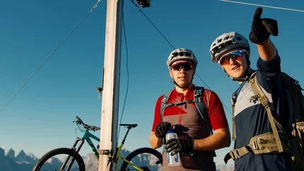 Zwei Radfahrer mit Helm und Rucksack am Gipfelkreuz mit Fahrrad im Hintergrund