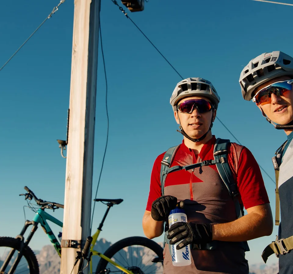 Mountainbike Urlaub für Anfänger
© David Karg Zwei Radfahrer mit Helm und Rucksack am Gipfelkreuz mit Fahrrad im Hintergrund