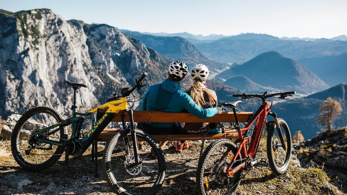 Blog © TVB Ausseerland Salzkammergut - Katrin Kerschbaumer Zwei Radfahrer schauen von Bank auf Berglandschaft mit zwei Mountainbikes