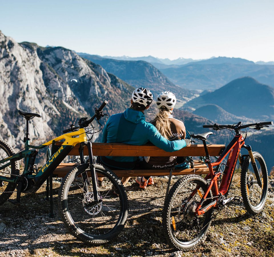 Zwei Radfahrer schauen von Bank auf Berglandschaft mit zwei Mountainbikes
