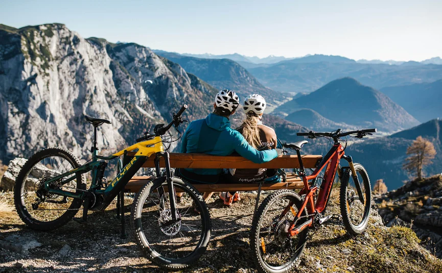 Two cyclists sitting on bench overlooking mountain landscape with two mountain bikes