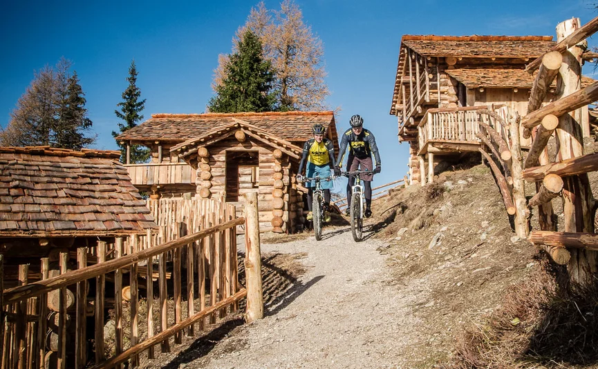 Zwei Mountainbiker fahren auf einem Weg neben Holzhütten bei klarem Himmel