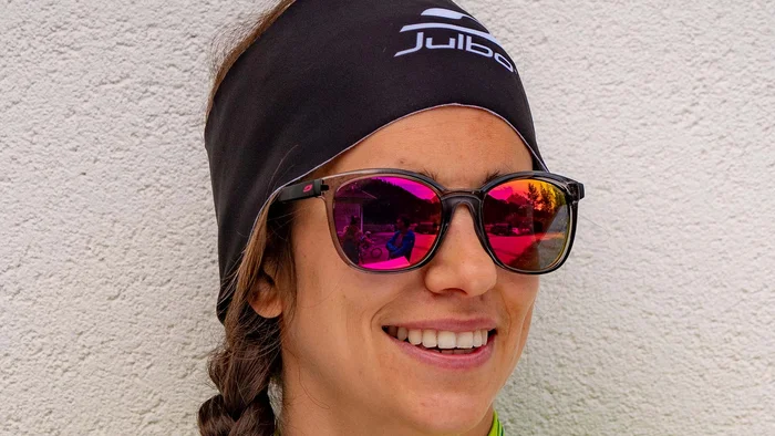Frau mit Sportbrille, Zopf und grün-blauem Bike School Trikot vor weißer Wand