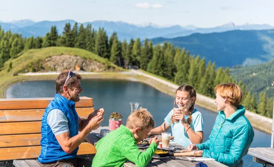 "Flotte Wadln" Wanderpauschale Familie isst zusammen draußen am See in den Bergen bei schönem Wetter
