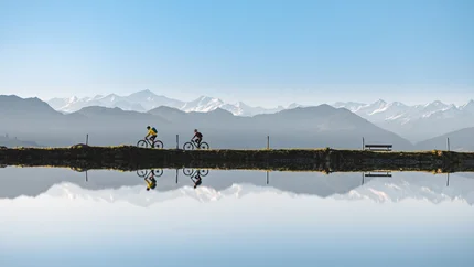 Zwei Radfahrer fahren am See mit Bergspiegelung und blauem Himmel