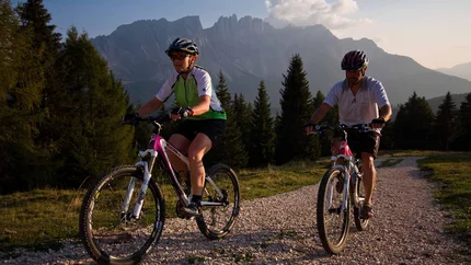 Twee personen mountainbiken op een grindpad met bergen en bomen op de achtergrond