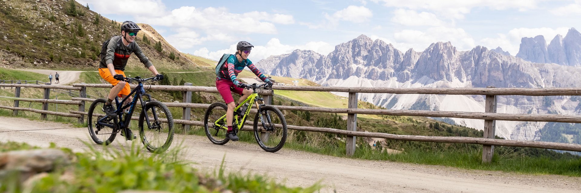 Zwei Radfahrer auf Bergpfad mit Holzzaun und Bergen im Hintergrund